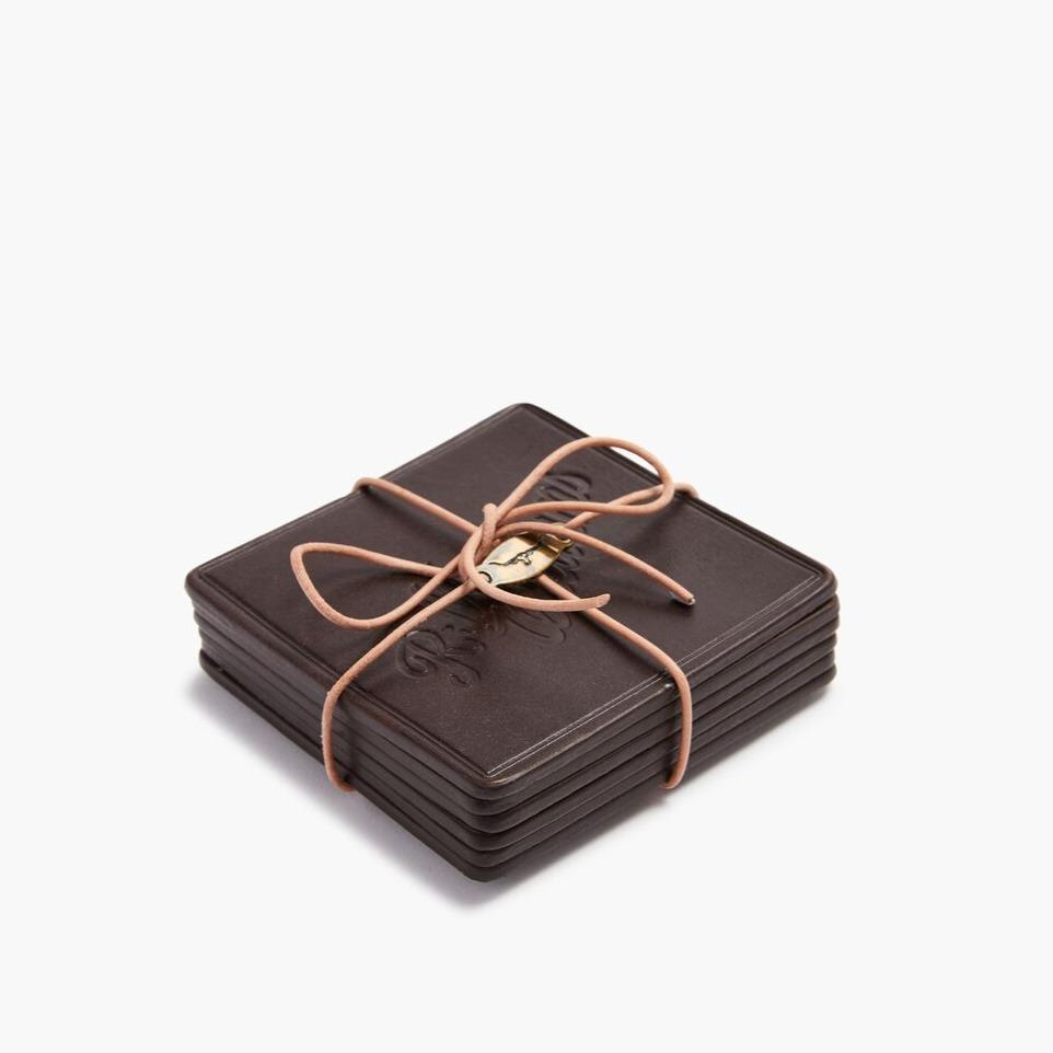 A set of six square, dark leather RM Williams coasters, tied with a light brown cord in a bow. The top coaster has an embossed design. Made with care in Australia, they add rich elegance to your table against a plain white backdrop.