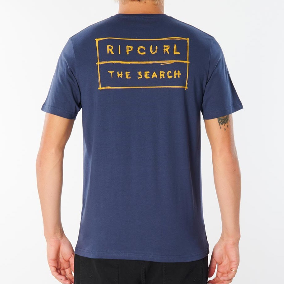 A person stands facing away from the camera, donning a dark blue Rip Curl 50/50 Scratch Tee with yellow text on the back that reads "RIPCURL THE SEARCH." The comfy tee, crafted from 100% cotton, reveals a tattoo on their right forearm and is set against a plain white background.