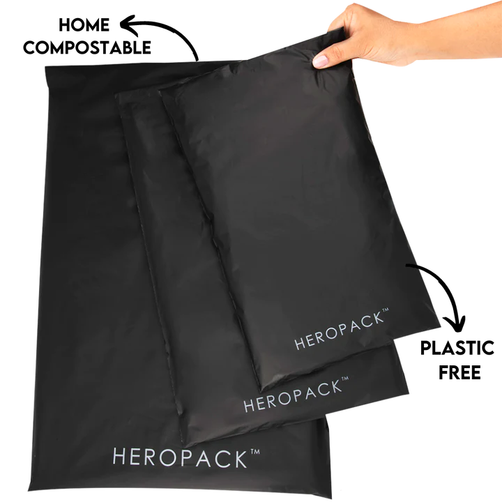 A hand is holding a black Compostable Eco Packaging mailing satchel. Two more of these bags, varying in size, are placed underneath the one being held. These sleek bags prominently feature "HEROPACK™" written in white at the bottom, highlighting their contribution to waste reduction and Greening Australia efforts.