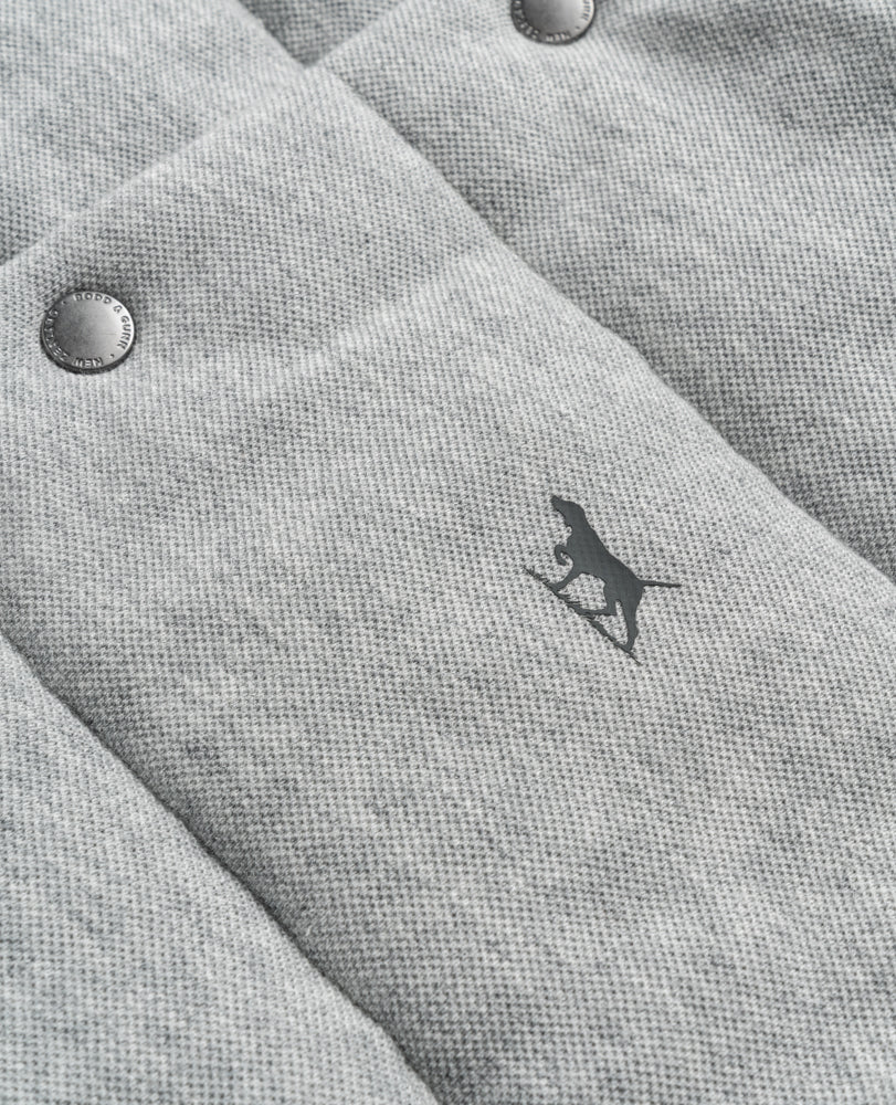The Rodd & Gunn Jackett Island Vest is a close-up of gray quilted fabric with a dog silhouette. It features two metallic buttons in the upper left corner and an adjustable waist for better fit. The vest offers soft fabric texture with high loft synthetic insulation for warmth and comfort, highlighted by a subtle pattern.