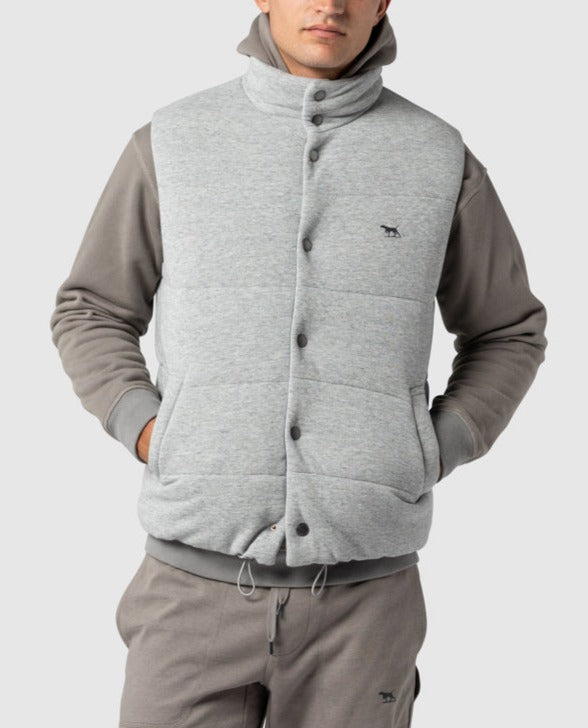 A man wears the Rodd & Gunn Jackett Island light gray padded vest with high loft synthetic insulation over a gray hoodie and matching pants. The vest features a small logo, button closure, adjustable waist, and his hands in pockets against a plain light gray background.