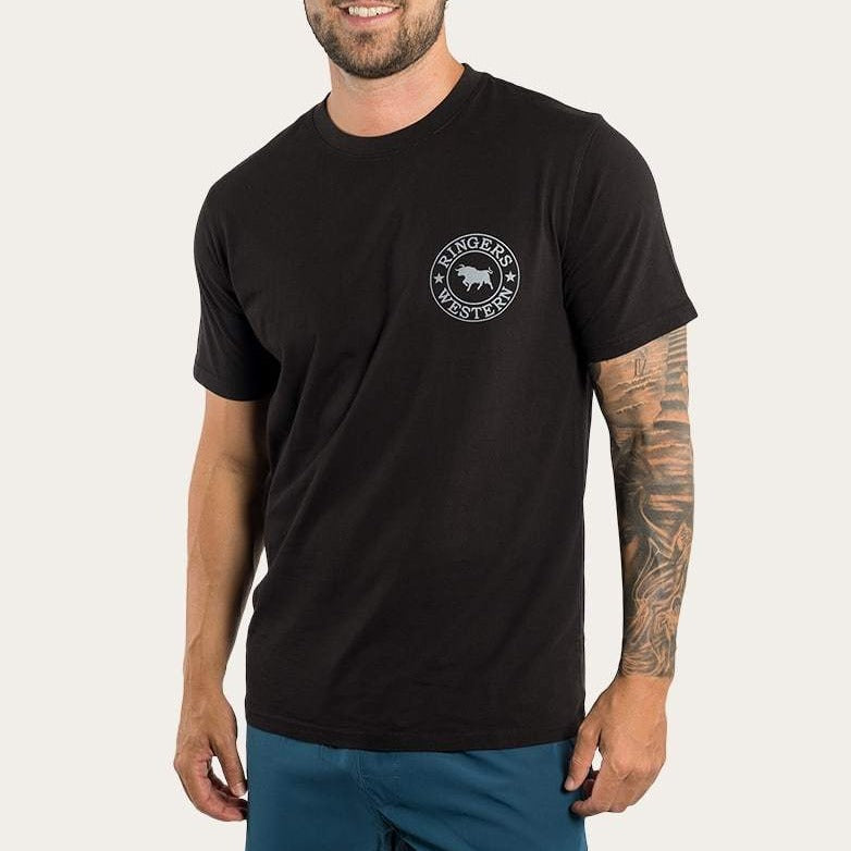 A person wearing the Ringers Western Signature Bull Classic T-Shirt, featuring 100% cotton and a small circular logo on the chest. The tattooed right arm is visible, paired with blue shorts. Set against a plain white background, this ensemble exudes a casual tee vibe.