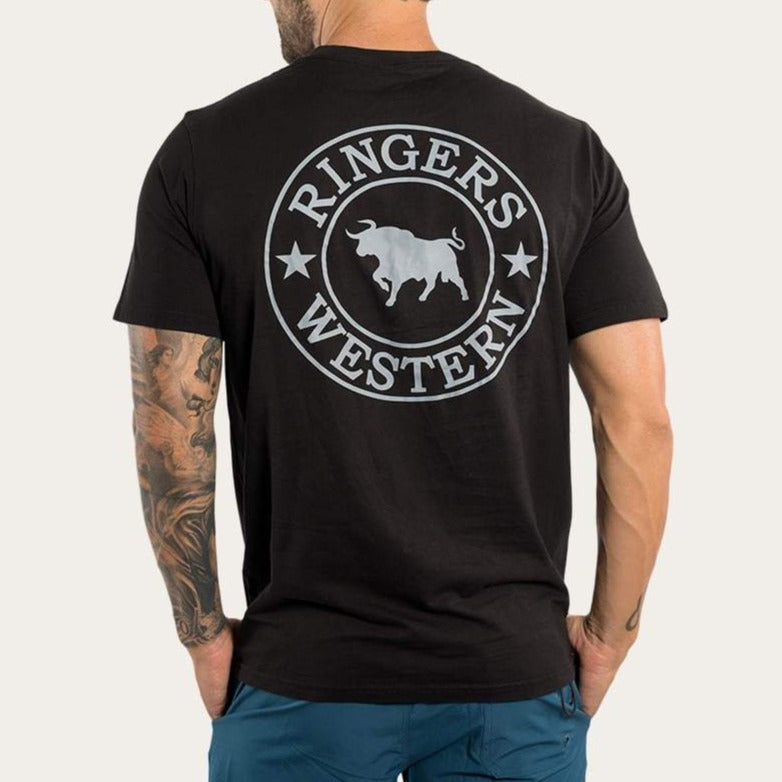 A man with tattooed arms is seen from behind wearing the black Ringers Western Signature Bull Classic T-Shirt, made of 100% cotton, featuring a circular "Ringers Western" logo with a bull and stars. He pairs it with blue pants for a casual look.