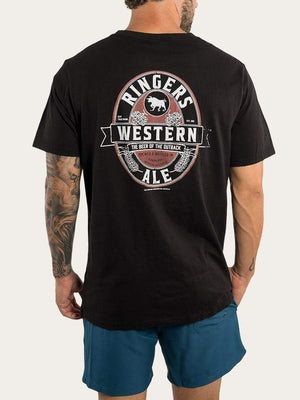 A man with a tattooed arm wears the Ringers Western Ale Loose Fit T-Shirt, made of 100% cotton, showcasing the logo and "The Beer of the Outback" text on the back. Paired with blue shorts, his look is ideal for relaxed days.