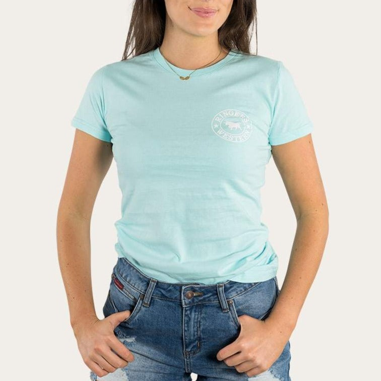 A person wearing a light blue Ringers Western Womens Signature Bull Classic Fitted T-Shirt with a small white circular logo on the left side of the chest and blue jeans, with hands in their pockets. The 100% cotton fabric ensures comfort. The background is plain and light-colored.