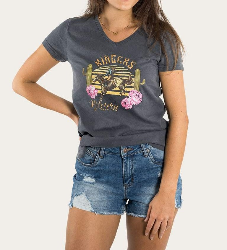 Wearing the Ringers Western Rodeo Women's Relaxed V-Neck Tee—a 100% cotton dark casual T-shirt with a western design of a cowboy, cacti, and flowers—paired with denim shorts, she effortlessly poses with one hand on her hip against a plain background.