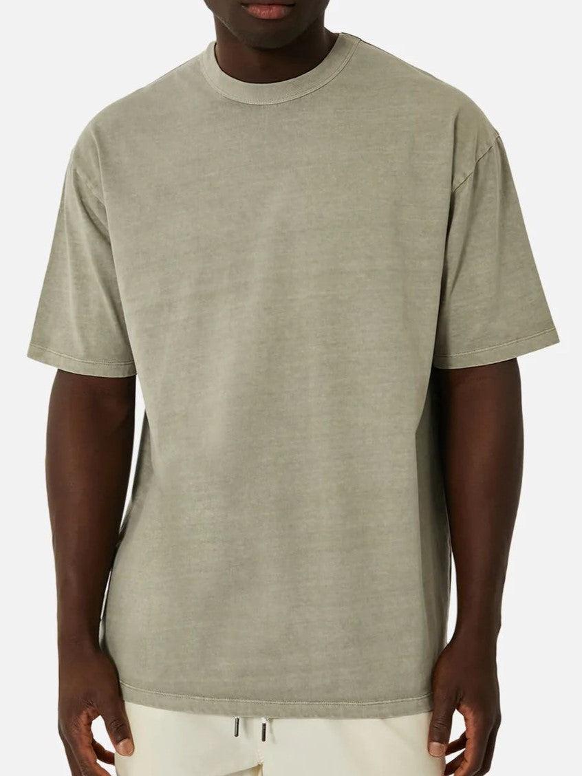 A person stands against a light gray background in the Industrie The Del Sur Tee, an oversized olive-green t-shirt with a crew neck and short sleeves, paired with light-colored pants; their hands rest by their sides.