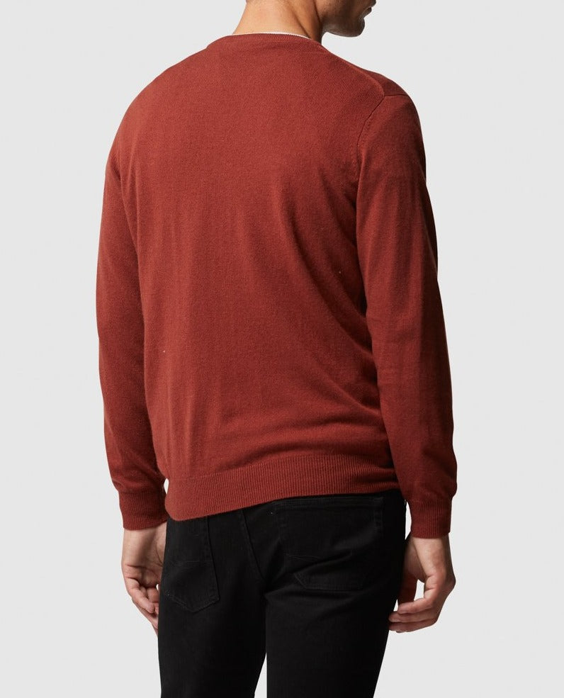 A person wearing a Rodd & Gunn Queenstown Sweater in rust and black pants stands with their back to the camera against a plain background, perfectly blending warmth and style for ideal travel layering.