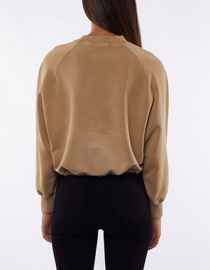 A person with long dark hair is shown from the back, wearing a beige Silent Theory Sleek Crew with raglan sleeves and black fitted pants. The cropped length sweatshirt perfectly complements the minimalist setting of the plain white background.