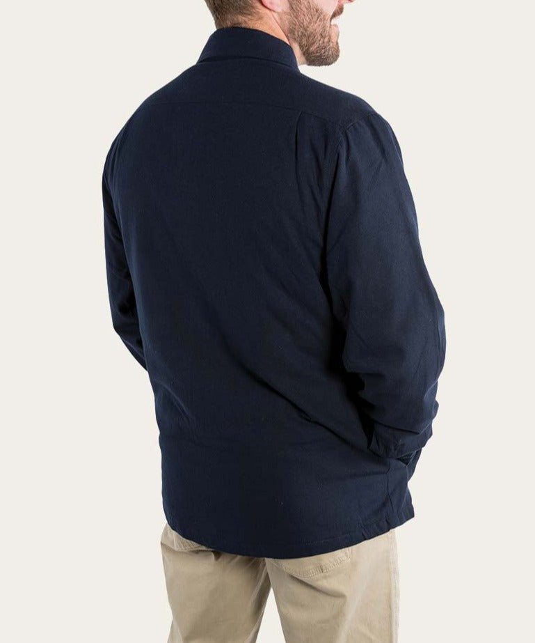 A man with short hair is seen from the back, wearing a dark blue Ringers Western Skipton Lumber Jacket and beige pants. He has his hands in the pockets of his comfortable jacket. The background is light and plain.