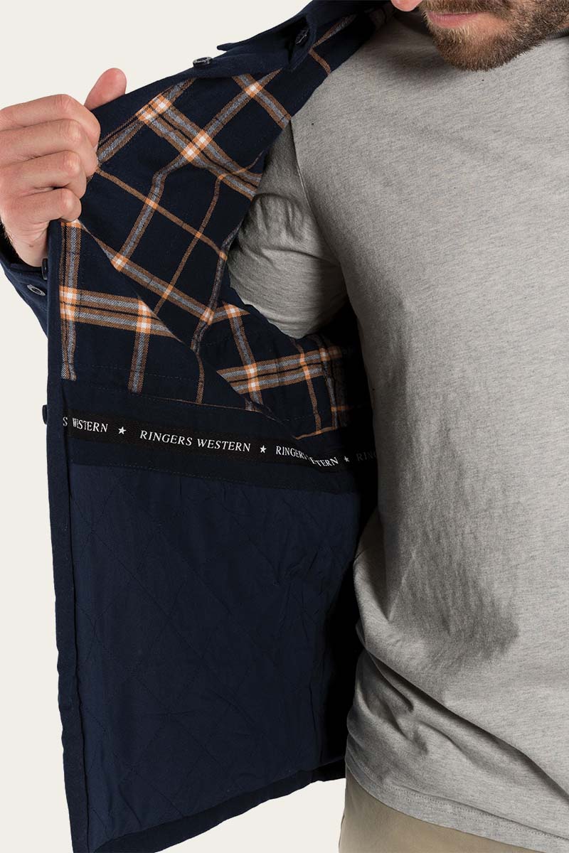 A person in a light grey t-shirt holds open a Ringers Western Skipton Lumber Jacket, revealing its plaid interior lining. The lining features a pattern of orange, white, and dark blue lines, with the brand name "Ringers Western" printed multiple times along the jacket's inner edge.
