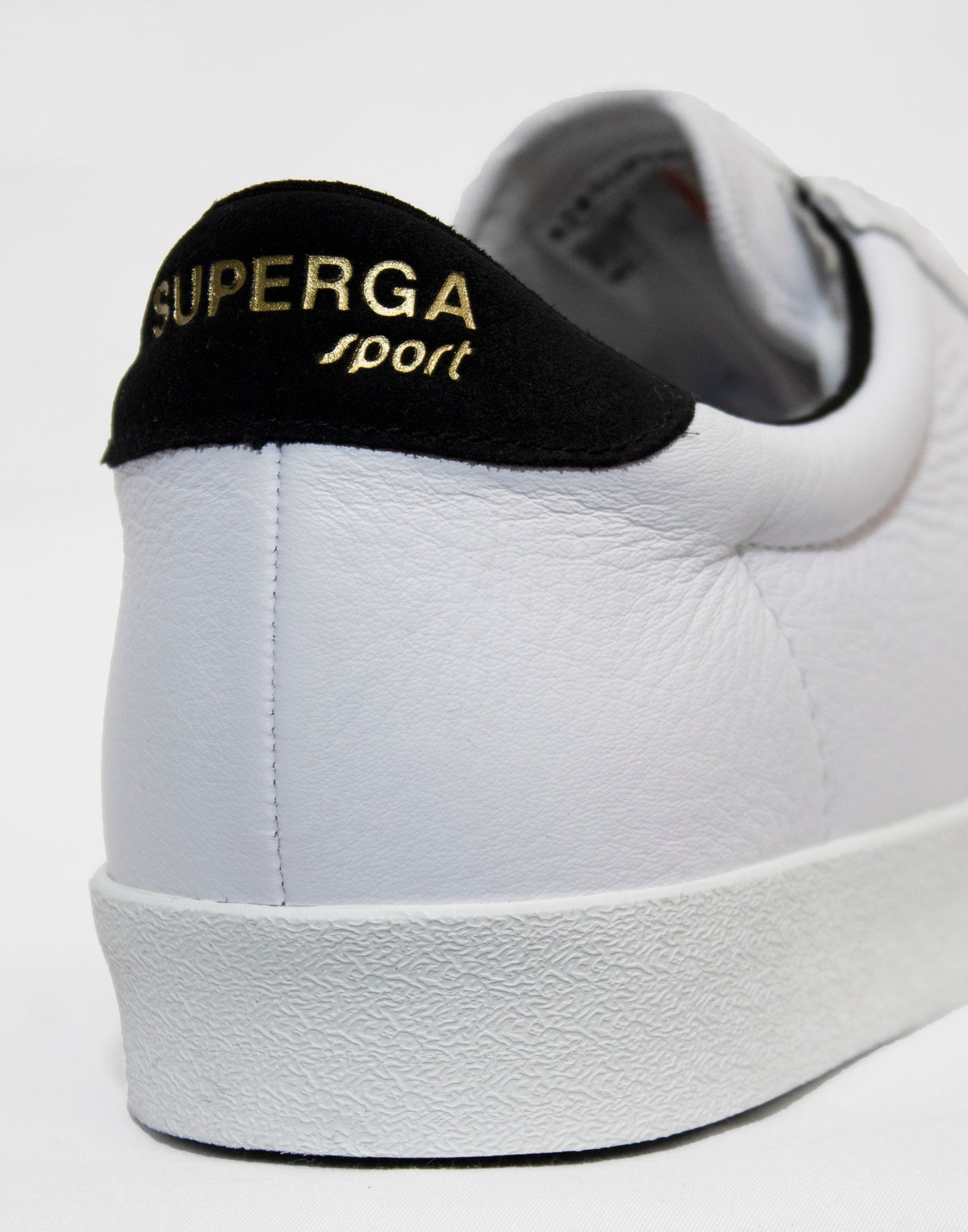 Close-up of a Superga Clubs Comfleasueu Sneaker, featuring a black heel tab with gold "SUPERGA sport" lettering. The shoe is on a white background, highlighting its textured leather surface and sleek design.