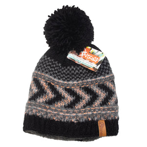 The Anticorp Ladies Arrow Lined Beanie is a knitted winter hat featuring a large black pom-pom on top. It showcases a black, gray, and white zigzag pattern with subtle orange accents and has a cozy fleece lining. The beanie comes with a colorful tag reading "Pistil" and has a small brown leather patch stitched to the brim.