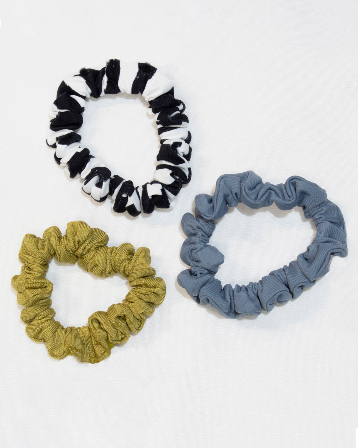 The Rip Curl 3 Pack Hair Scrunchie set is showcased on a white background. The top scrunchie features black and white stripes, the bottom left scrunchie is yellow-green, and the bottom right scrunchie is light blue. Each scrunchie combines elastane and polyamide for a soft, stretchy feel.