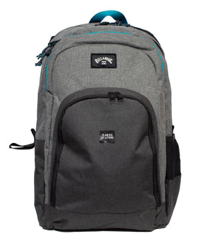 The Billabong Command Backpack is gray and black with a front zipper pocket, mesh side pocket, teal-accented top handle, dedicated laptop compartment, and a front label reading "9.6 kg, 27L.