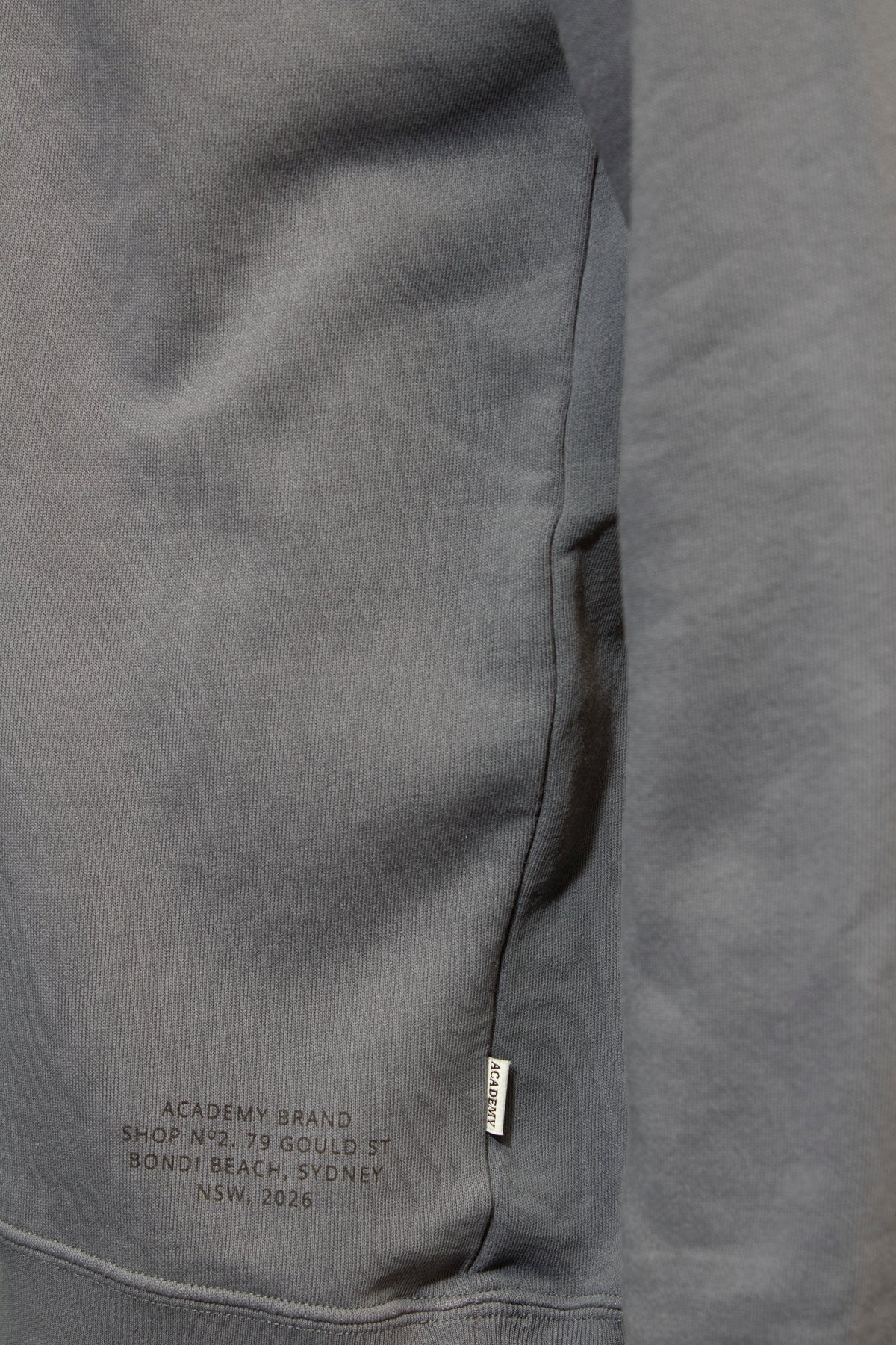Close-up of a grey Academy Brand Bondi 1/2 Zip Sweat showing a small tag on the seam and printed text: "ACADEMY BRAND, SHOP 1, NO. 79 GOULD ST, BONDI BEACH, SYDNEY, NSW 2026.