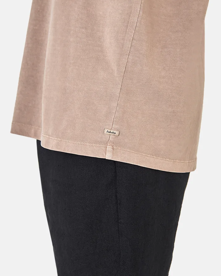Close-up of a person wearing Industrie The Del Sur Tee, a beige casual tee and black pants. The focus is on the shirt's side, showing a small fabric tag with text. Made from 100% cotton, the image captures its texture and relaxed fit.