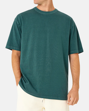 A person wearing an oversized acid wash teal T-shirt and light-colored pants stands with one hand in their pocket against a plain background. The Industrie The Del Sur Tee offers a relaxed fit with short sleeves, combining style and comfort seamlessly.