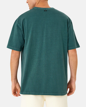 Wearing the Industrie The Del Sur Tee, an individual in a teal oversized T-shirt stands with their back to the camera, paired with light-colored shorts against a neutral gray background.