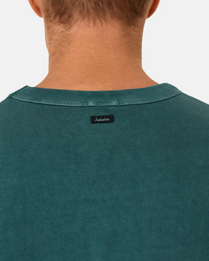 A person is seen from the back, wearing the Industrie The Del Sur Tee, an oversized teal shirt with an acid wash. A black tag near the collar displays "Indestructible" in white script against a plain white background, accentuating its contemporary style.