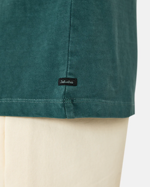 A close-up of the hem of a dark teal, oversized fit shirt with a small black "Industrie" label. The comfortable Industrie The Del Sur Tee is paired with light beige pants against a plain white background.