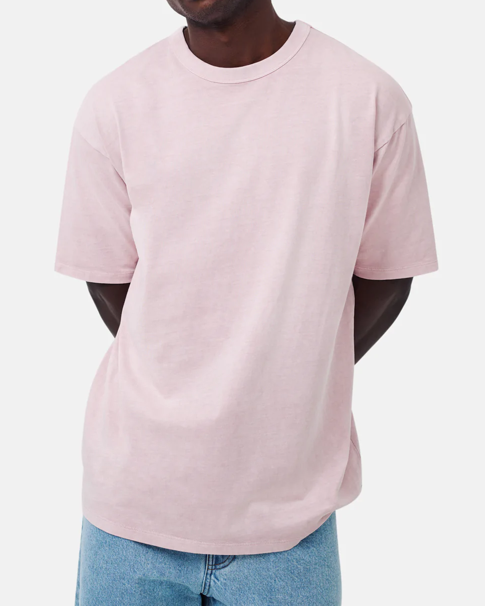 A person models the Industrie The Del Sur Tee in pastel pink, made of 100% cotton, paired with light blue jeans against a plain white background. The comfortable tee boasts an oversized fit, with the head cropped out of view and arms positioned behind their back.