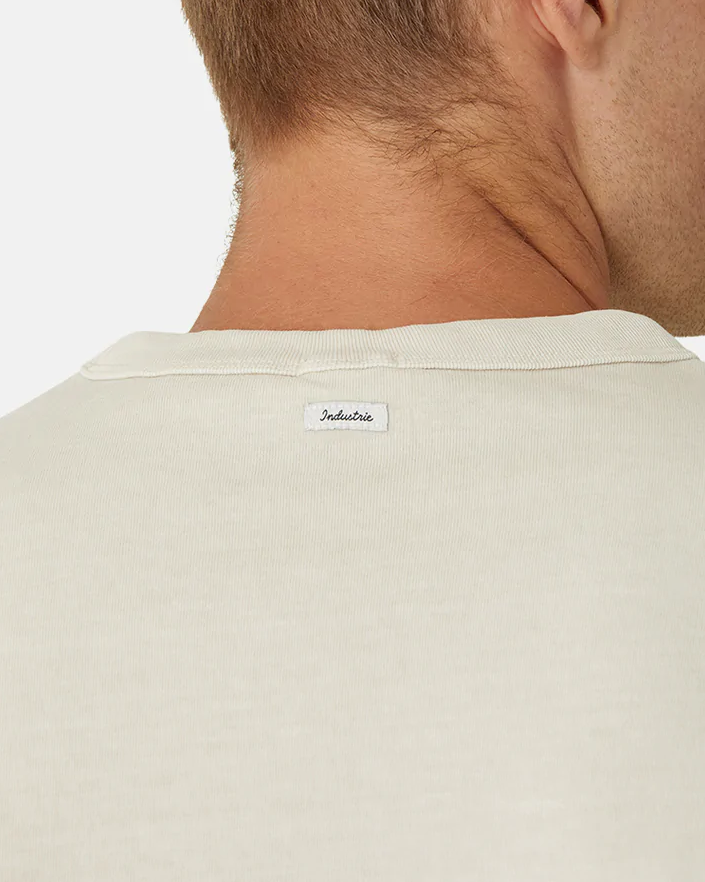 A close-up shows the back of a person's neck wearing an oversized, beige tee labeled "Industrie The Del Sur Tee." Made from 100% cotton, the comfortable shirt reveals their short hair above the collar.