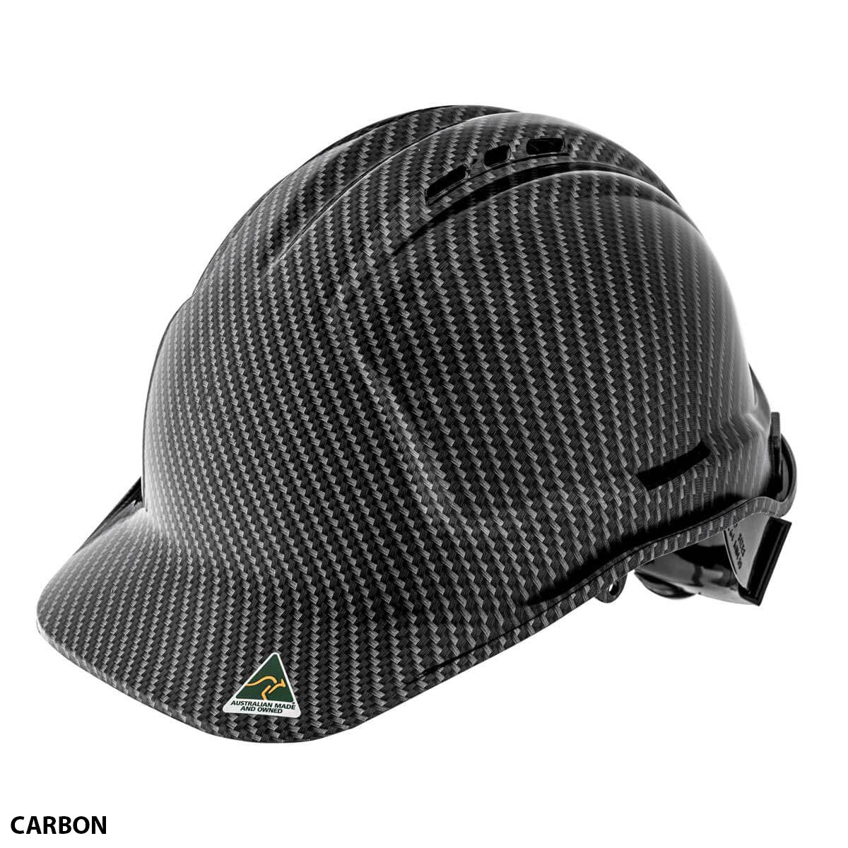The SSA Custom Hydro Dip Hard Hat Vented is a carbon fiber-look safety helmet with a sleek design and an adjustable strap. It is AS/NZS 1801:1997 certified and features a small logo of a green triangle with a yellow border and a mountain inside, ventilation slots on the top for airflow, and the word "CARBON" in the bottom left corner.