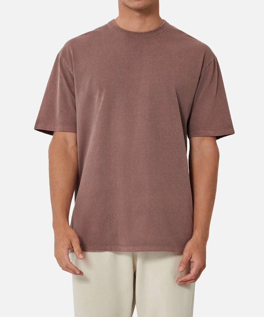 A person in an oversized fit, Industrie The Del Sur Tee, a plain brown T-shirt with short sleeves and a round neckline, paired with off-white pants, stands against a neutral background. The person's face is not visible.