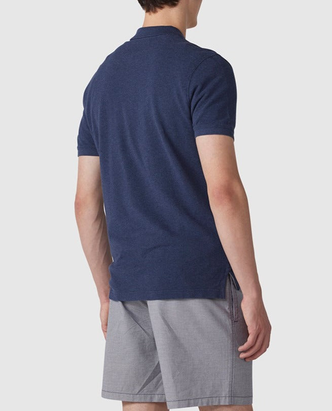 A person is standing with their back to the camera, wearing the Rodd & Gunn The Gunn Polo in navy blue, a modern fit, short-sleeve shirt made from Australian Cotton, paired with gray shorts. The background is plain and light-colored.