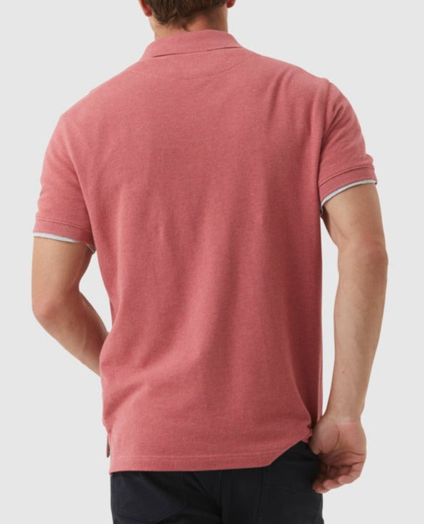 A man is shown from the back wearing a short-sleeved Rodd & Gunn The Gunn Polo in pink, crafted from Australian Cotton, along with dark pants. He is holding the bottom hem of his comfortable polo with one hand, adjusting it slightly. The background is plain and light grey.