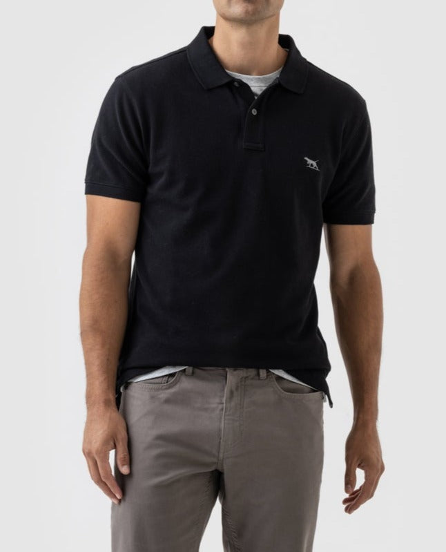 A person is wearing a black Rodd & Gunn The Gunn Polo and gray pants. The polo shirt, made from Australian Cotton, features a small Rodd & Gunn logo embroidered on the left chest. They are standing against a plain, light gray background. The person’s head is not visible in the image.