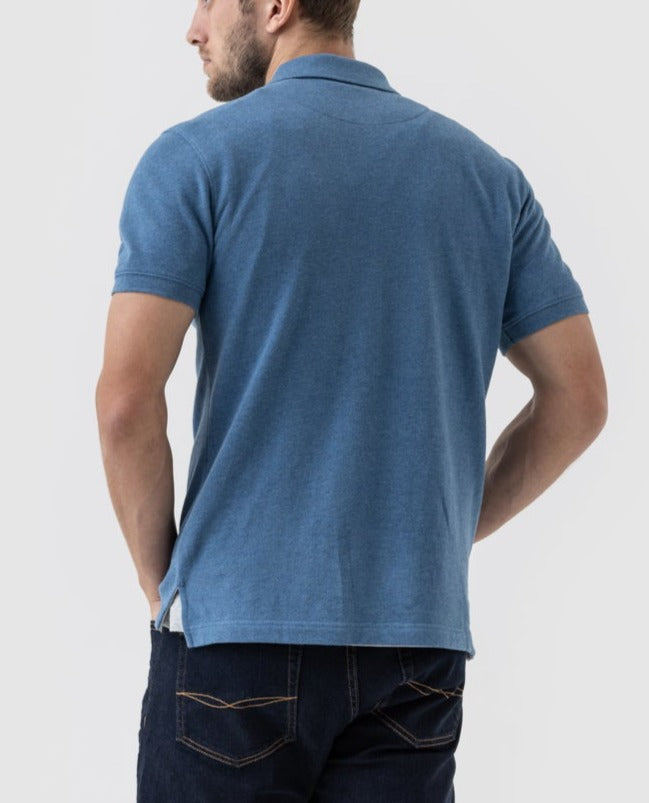 A person stands facing away, wearing the Rodd & Gunn The Gunn Polo in blue short sleeves and dark blue jeans. With hands placed on their hips, they are set against a plain light grey background.