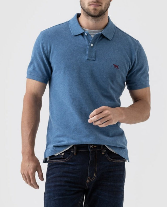 A man is shown from mid-thigh up against a plain background, dressed in a short-sleeved, blue Rodd & Gunn The Gunn Polo shirt and dark jeans. The Australian Cotton polo features a small red emblem on the left side of his chest. His right arm hangs relaxed by his side, while his left hand is raised slightly.
