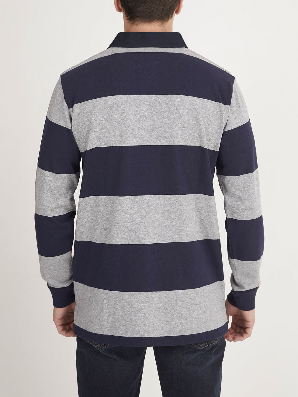 A person is standing with their back to the camera, wearing a RM Williams Tweedale Rugby shirt made of 100% cotton, featuring wide horizontal stripes in alternating navy blue and light gray colors. They are also wearing dark blue jeans. The background is plain white.