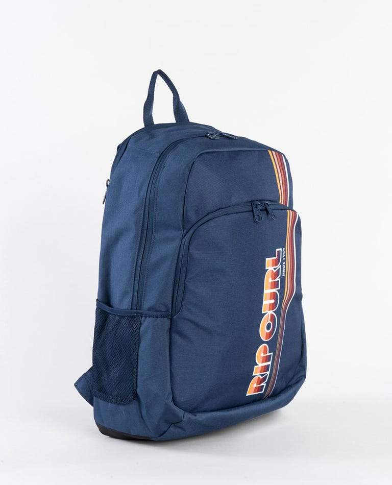 A Rip Curl Ozone 30L Backpack in navy blue comes equipped with multiple compartments and zippers. It features a front pocket adorned with the Rip Curl logo in bold white and orange letters set against a stripe-patterned background. The side boasts a mesh pocket, adjustable straps, an insulated cooler compartment, and the top is finished with a carrying handle.