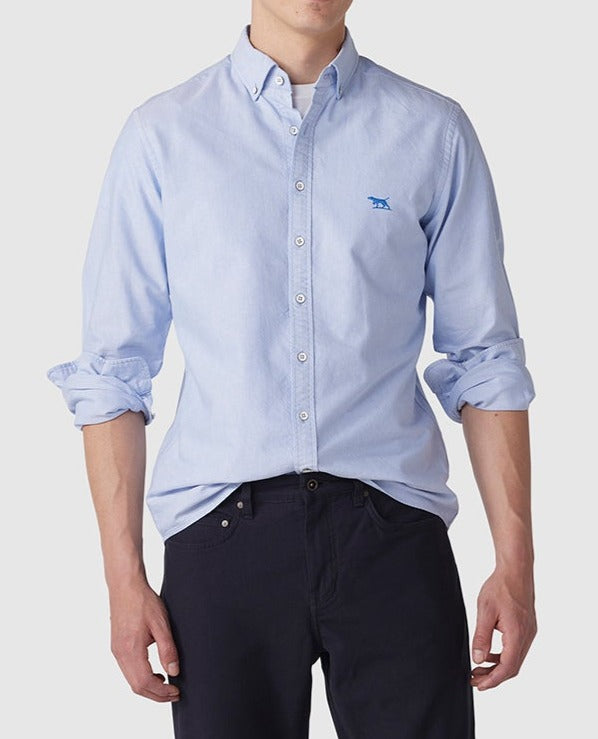 A person wears a light blue Rodd & Gunn Gunn Oxford Sports Fit Shirt made of soft cotton, featuring rolled-up sleeves and a discreet blue logo on the chest, paired with dark pants. The shirt is slightly tucked in at the front against a plain white background.