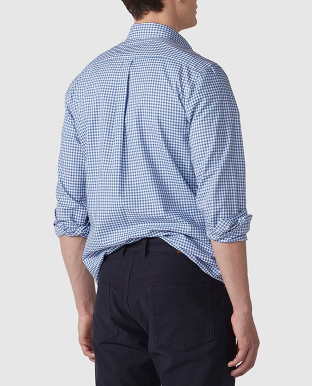 A man is photographed from behind, wearing the stylish Rodd & Gunn Gunn Check Oxford Sports Fit Shirt, which has a blue and white-checkered pattern. The shirt, made of 100% cotton, has its sleeves rolled up and is loosely tucked in at the back. He pairs it with dark-colored pants against a plain, light grey background.