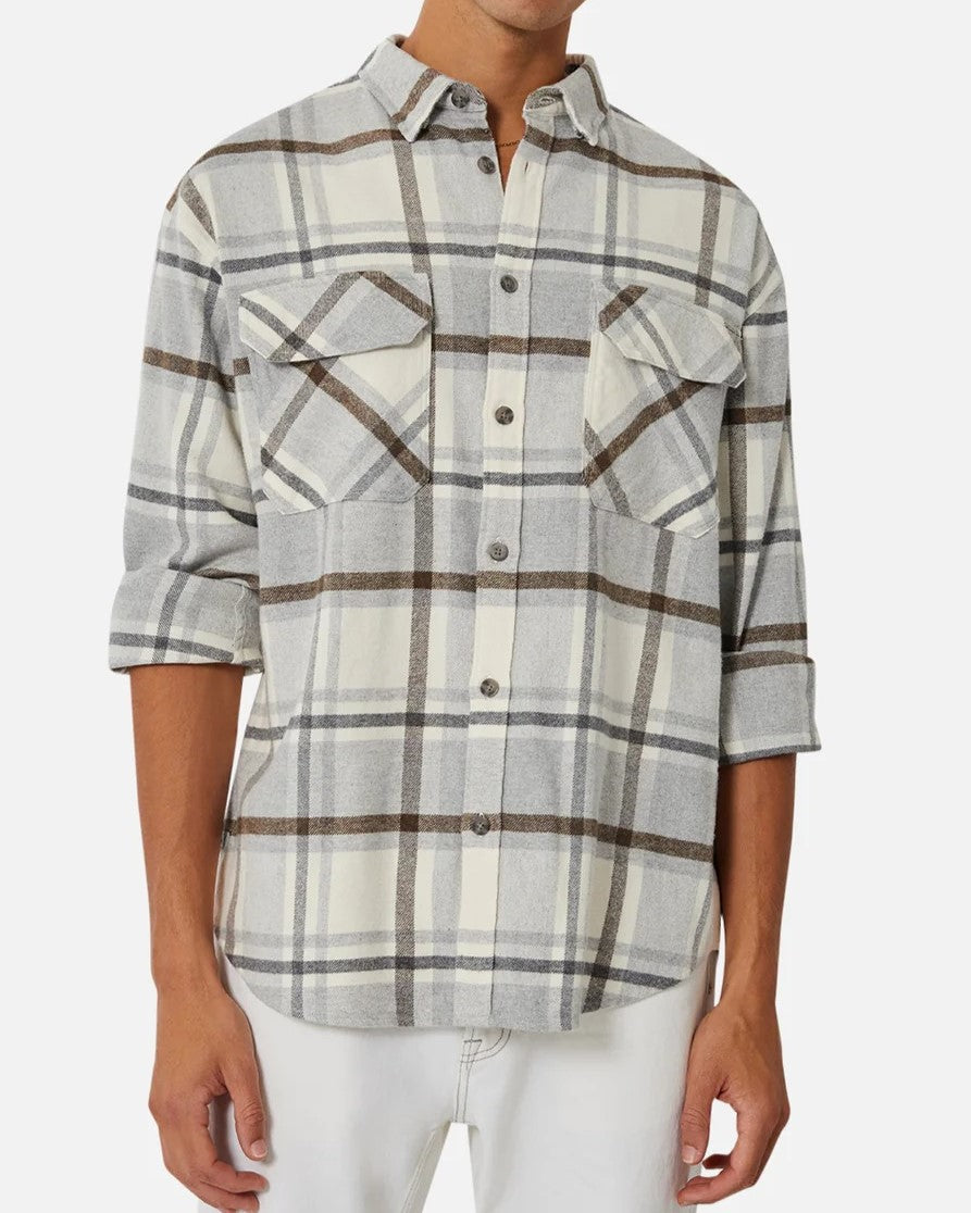 A person is wearing the Industrie The Elmhurst Shirt, a gray and white plaid relaxed fit button-up with rolled sleeves. Made from brushed cotton, it features two chest pockets and is paired with white pants. The person's head isn't visible in the image.
