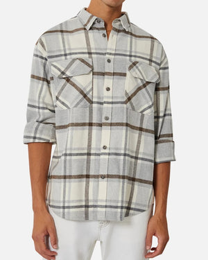 A person is wearing the Industrie The Elmhurst Shirt, a gray and white plaid relaxed fit button-up with rolled sleeves. Made from brushed cotton, it features two chest pockets and is paired with white pants. The person's head isn't visible in the image.