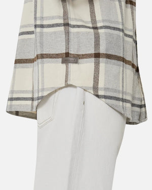 Close-up of a person in the Industrie The Elmhurst Shirt, a beige and brown plaid overshirt made from brushed cotton, paired with light pants. A small label is on the shirt's bottom. A plain white background emphasizes its relaxed fit and casual elegance.