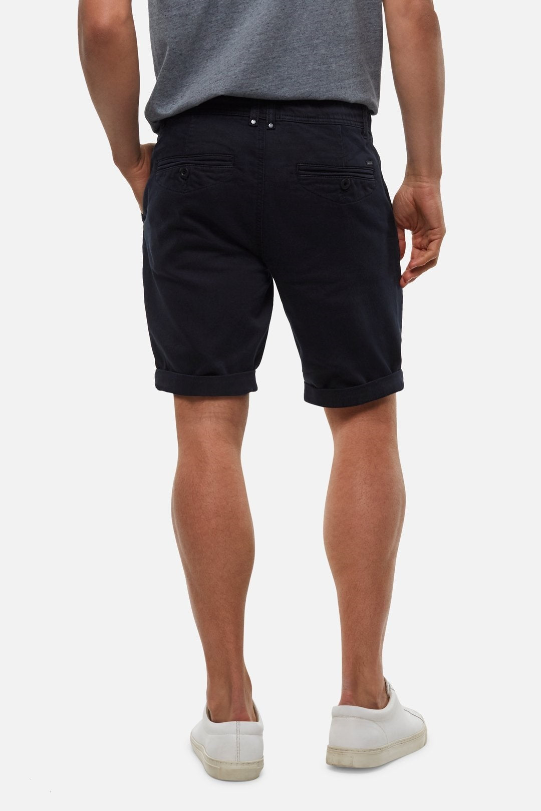 A person wearing a gray T-shirt, dark navy **Industrie The Drifter Cuba Shorts**, and white casual shoes is standing with their back to the camera, against a white background. The modern slim fit shorts feature two back pockets and are rolled up slightly at the hems.