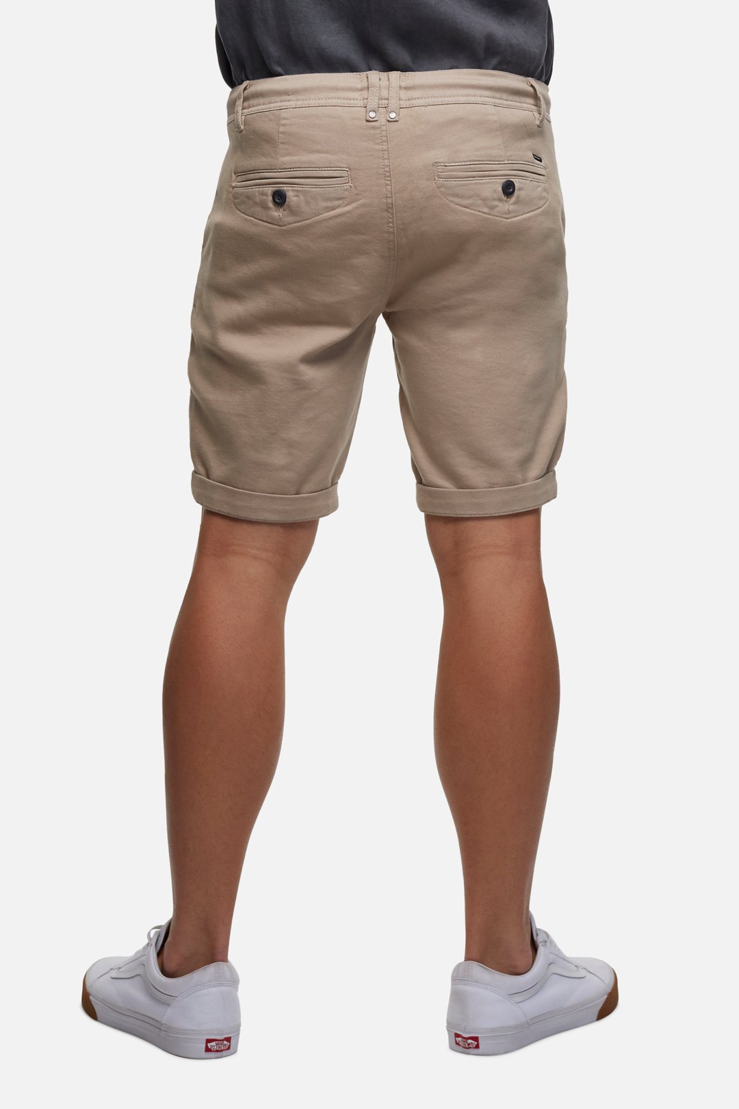 The image shows the back view of a person wearing Industrie The Drifter Cuba Short in a modern slim fit beige style paired with white sneakers. The comfortable shorts feature two back pockets with buttons. The person is also wearing a dark-colored shirt tucked into the shorts, set against a white background.