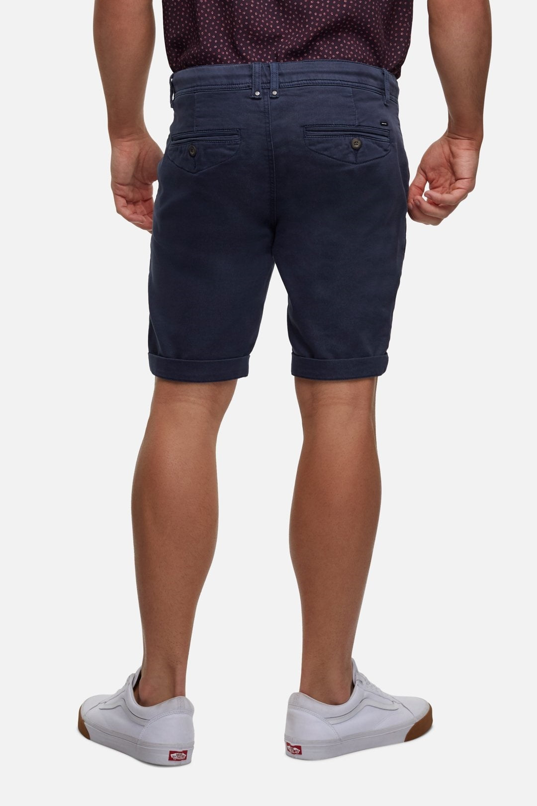 Rear view of a person wearing a dark maroon short-sleeved shirt tucked into Industrie The Drifter Cuba Short, modern slim fit navy blue casual shorts with rolled-up hems. The person is also wearing white sneakers with brown soles. The background is plain white.