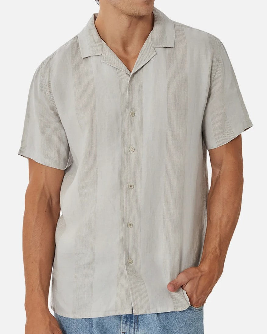 A person wears the Industrie The Kanaki Short Sleeve Shirt in light gray, featuring subtle vertical stripes, paired with light blue jeans and stands with one hand in their pocket against a plain background.