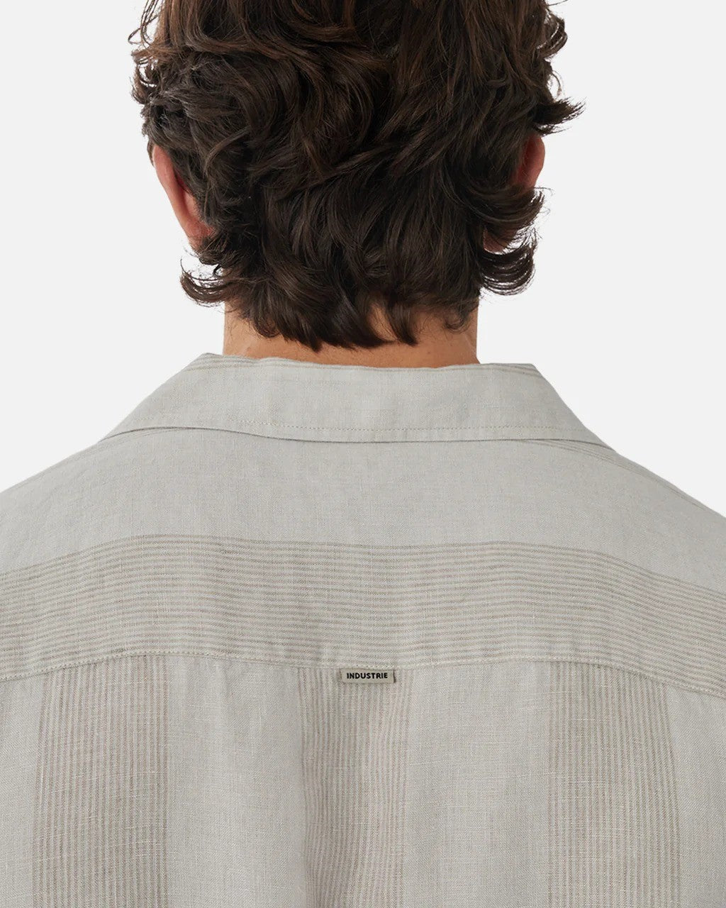 From behind, a person with wavy brown hair wears the Industrie The Kanaki Short Sleeve Shirt in light gray linen with vertical stripes. It features a "INDUSTRIE" label on the upper back. The background is plain white.