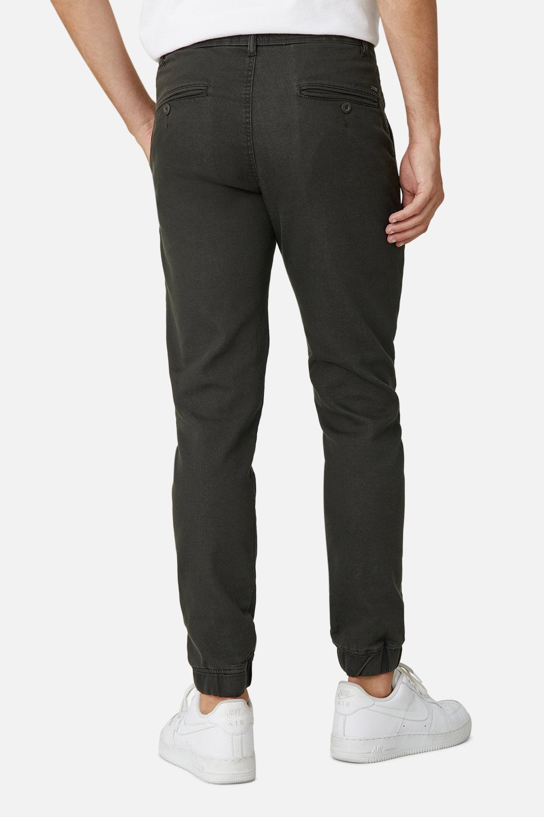 A person stands against a white background, wearing Industrie The Drifter Chino Pant in dark green with an elastic waist and tapered ankles, paired with white sneakers. One hand is casually placed in a back pocket.