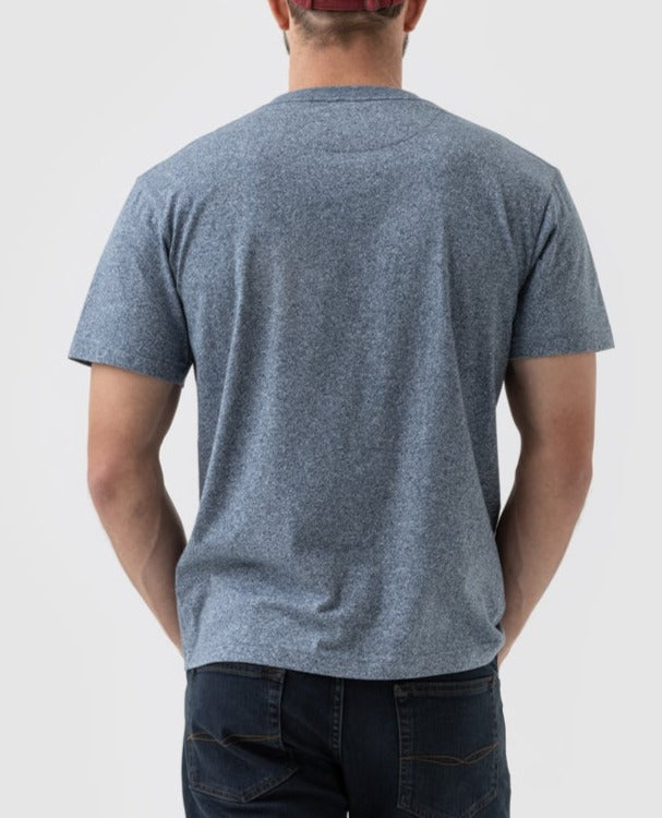 A person wearing a Rodd & Gunn The Gunn T-Shirt in heather blue, paired with dark blue jeans and a red cap, stands with their back facing the camera against a light grey plain background.
