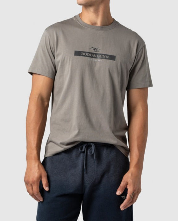 A man wears a gray Rodd & Gunn Cumulus Sports Fit Tee, made of premium cotton and featuring "RODD & GUNN" on the front. The comfortable casual tee pairs perfectly with his navy blue shorts as he stands against a plain white background.