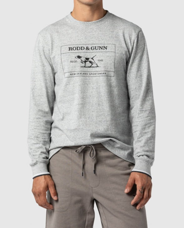 A person wears a Rodd & Gunn Bonny Glen Sports Fit T-Shirt in light gray, crafted from 100% cotton with a black dog logo and "New Zealand Sportswear" text. Paired with taupe pants, their stylish outfit pops against the plain white background.